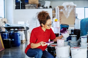 美しい白人の女性従業員が、色とりどりのバケツの隣にしゃがみ込み、印刷のために色を混ぜています。印刷所のインテリア。