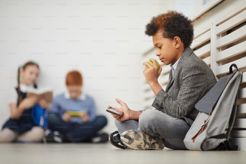 African schoolboy resting on the floor eating fruit and using his mobile phone with his classmates in the background