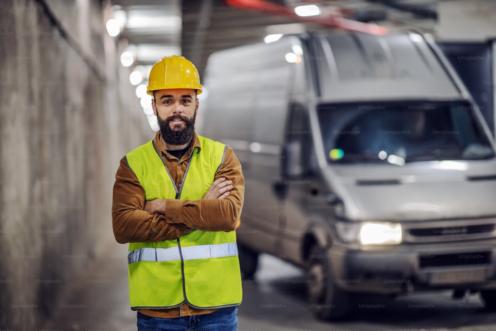 Young handsome bearded smiling supervisor in vest, with safety helmet on head standing in underground parking lot in construction process with arms crossed. In background is pickup truck.