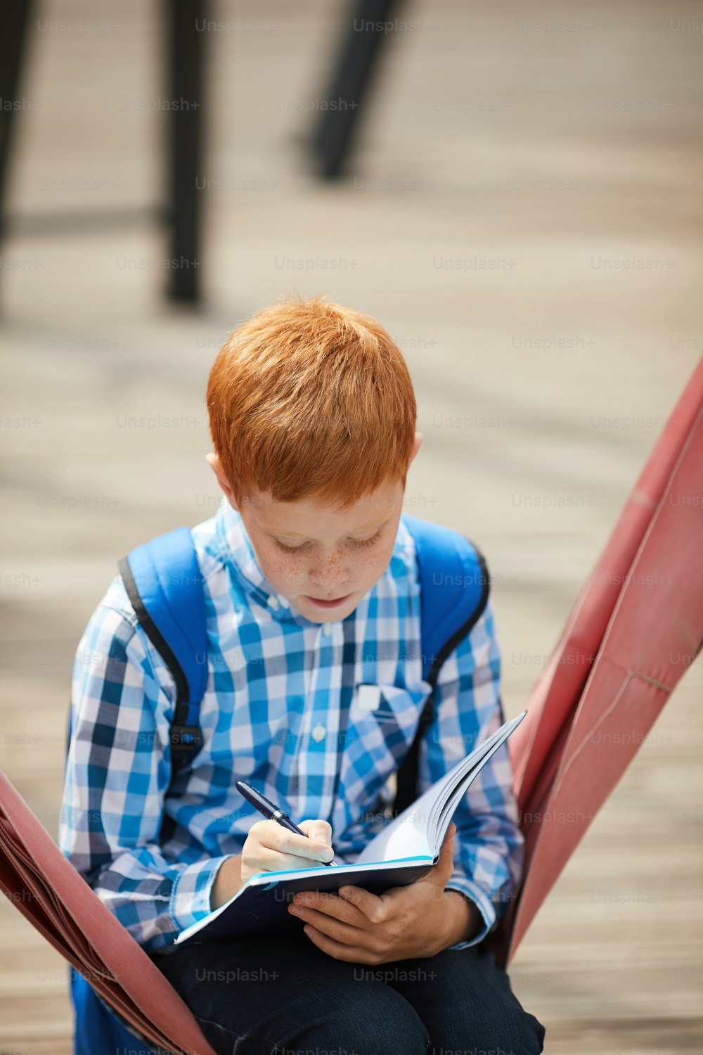 Red haired schoolboy sitting on the swing and making notes in his notebook he doing his homework outdoors