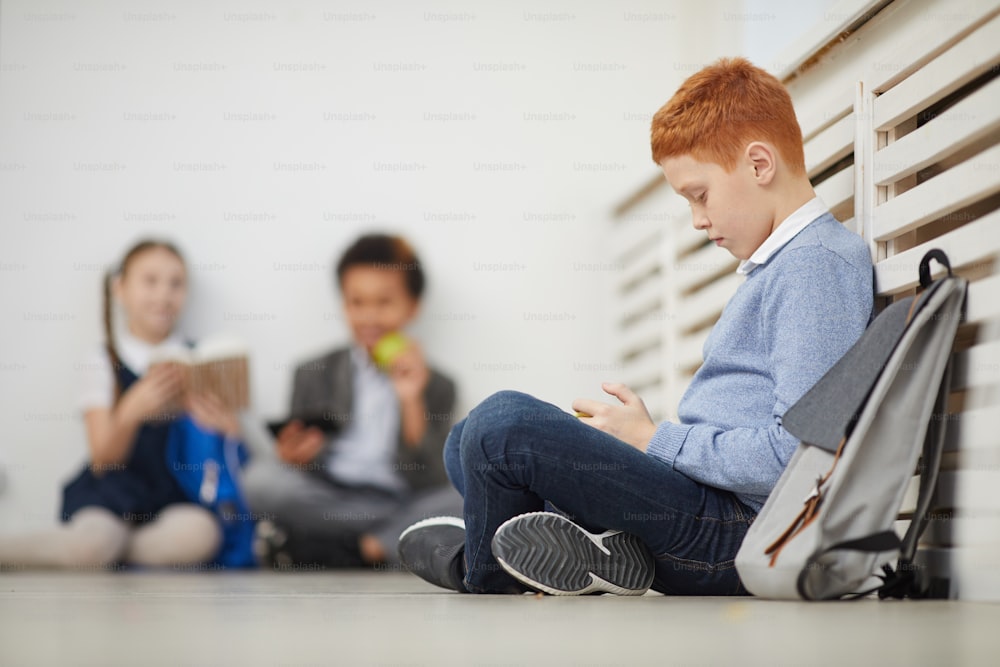 Red haired schoolboy sitting on the floor and playing in mobile phone during a break at school with other children in the background