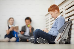 Red haired schoolboy sitting on the floor and playing in mobile phone during a break at school with other children in the background