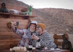 Two elderly people in love look at the cellphone for a selfie. Food and drink on the table with salami and red wine. Mountain in background