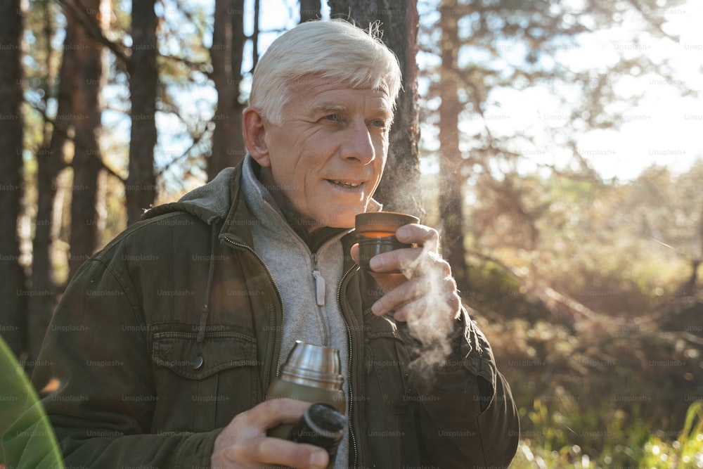 회색 머리의 남자가 향기로운 음료를 즐기며 아침 가을 숲에서 차분한 느낌을 받으며 뜨거운 차와 함께 컵을 들고 있습니다.