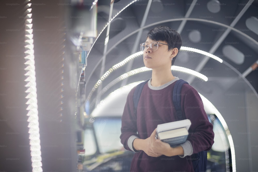 Asian student in eyeglasses holding books and looking at bookshelf while standing at modern library
