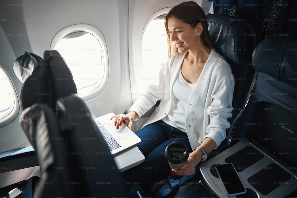 Fröhliche kaukasische Frau sitzt im Flugzeug und lächelt, während sie an einem Laptop arbeitet und Kaffee trinkt