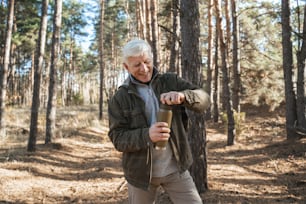 Cheerful old man is opening thermos while preparing pouring hot tea while traveling in wild nature and standing at the forest. Hiking concept. Stock photo