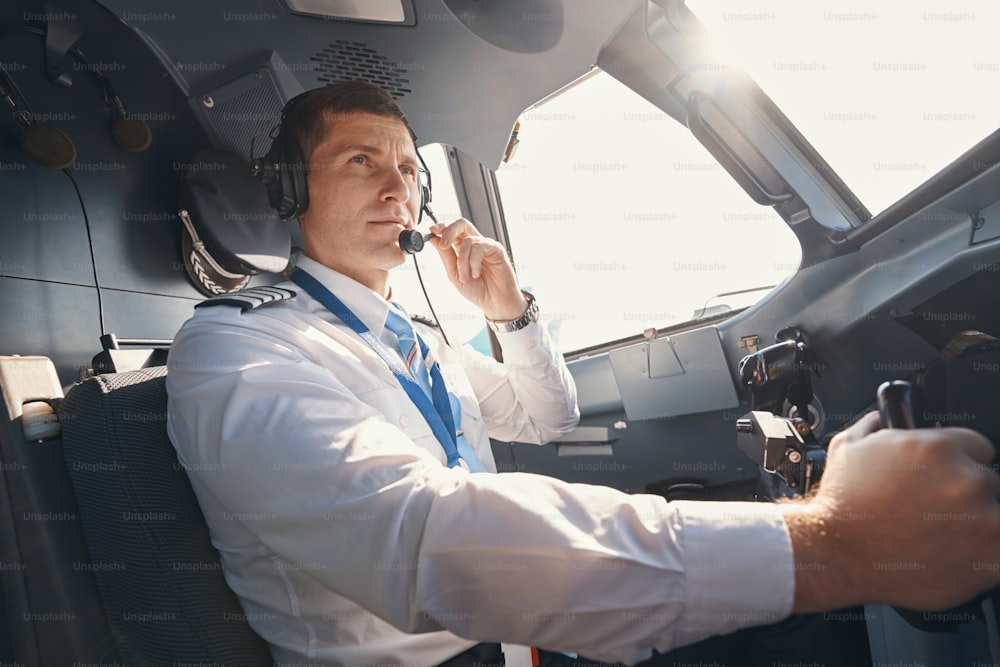 Piloto caucásico en camisa blanca hablando por micrófono de auriculares desde la cabina del avión