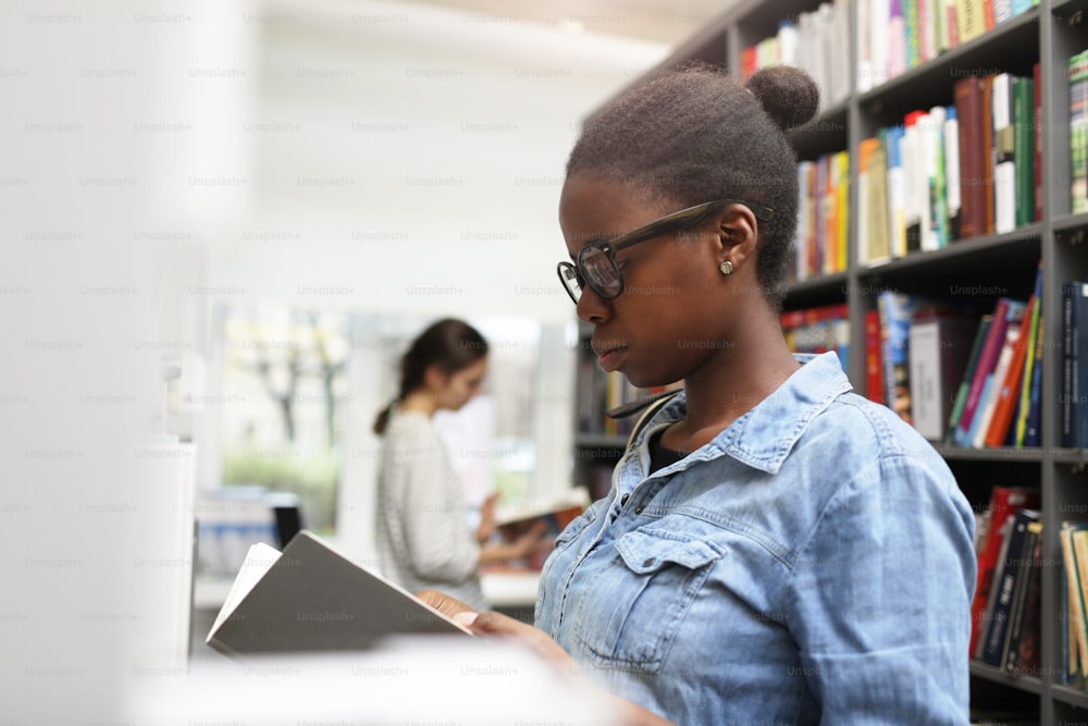 Serious African woman wearing eyeglasses examining the book she searching for the book in the library