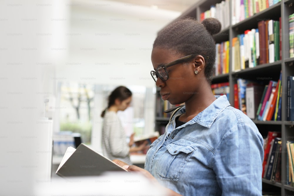 Donna africana seria che indossa occhiali esaminando il libro che cerca il libro in biblioteca