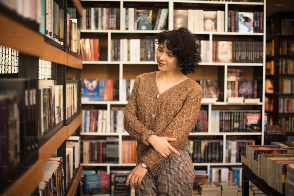 Mujer joven sonriente de pie en la biblioteca y mirando libros.