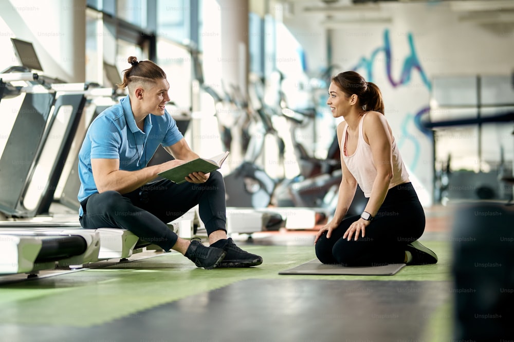 Happy male coach communicating with sportswoman who is taking a break from exercising in a gym.