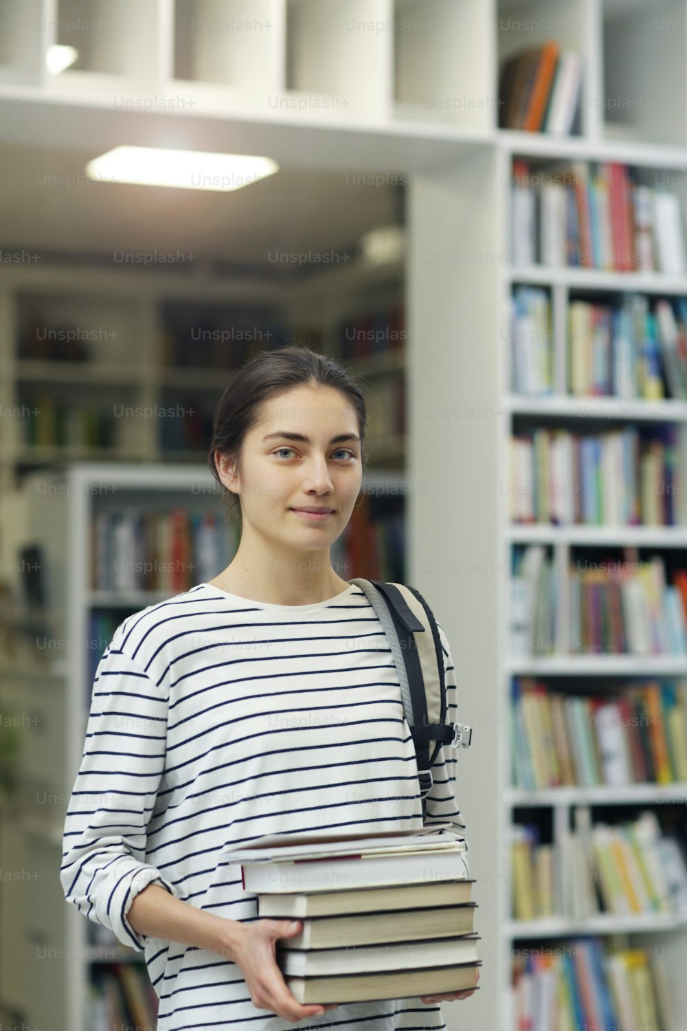 Portrait of young beautiful woman holding books and looking at camera while standing in the library