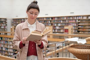 Mujer joven seria examinando el libro mientras está de pie en la gran biblioteca