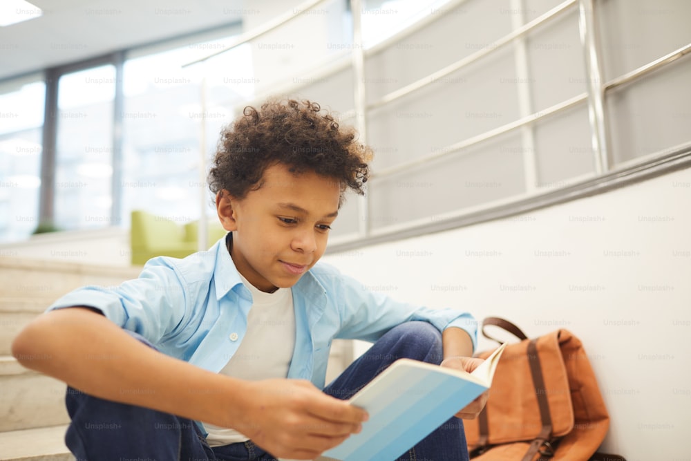 Horizontal medium portrait of curious African American boy reading interesting book sitting alone on stairs, copy space