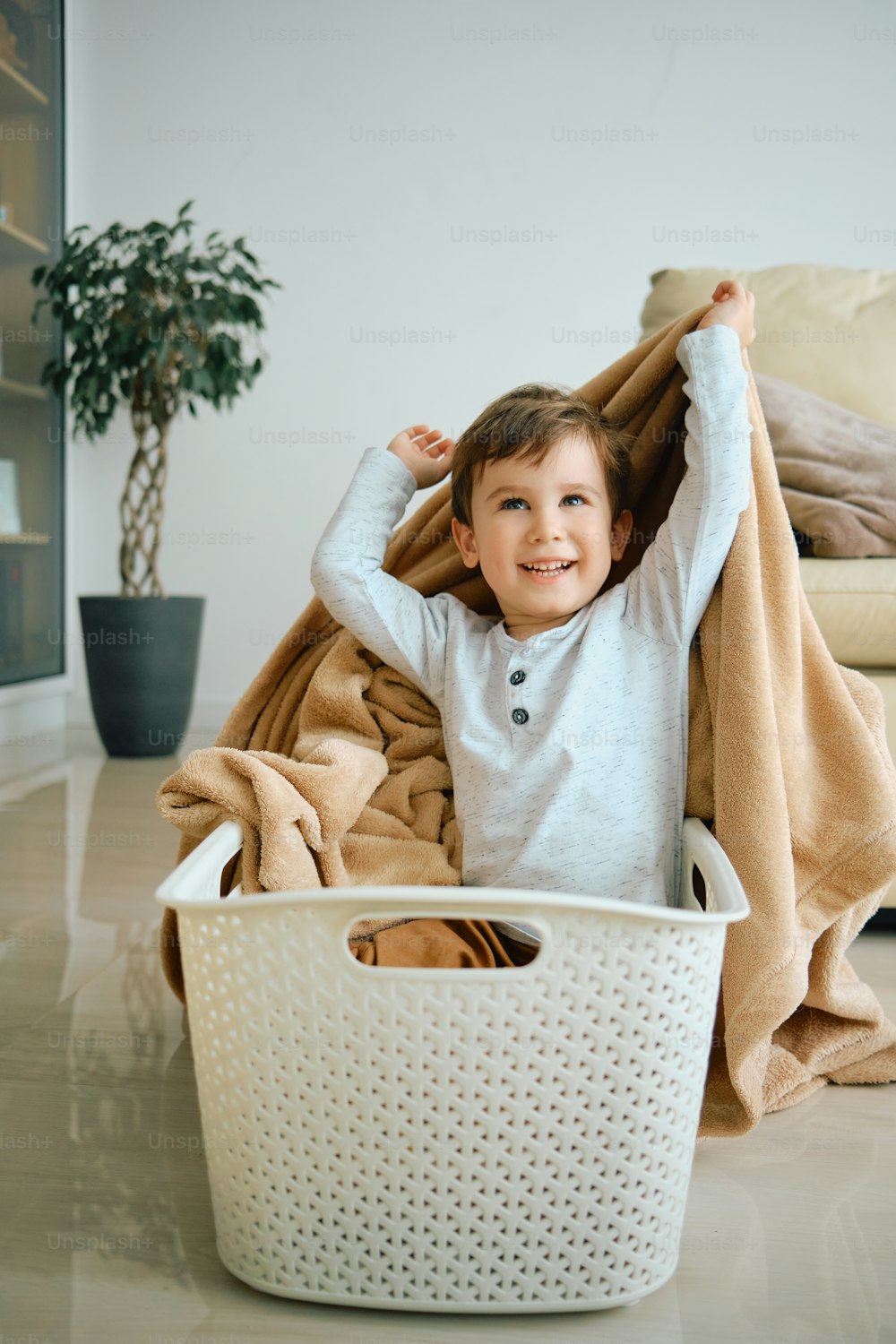 Happy little boy sitting in laundry basket and playing with a blanket at home.