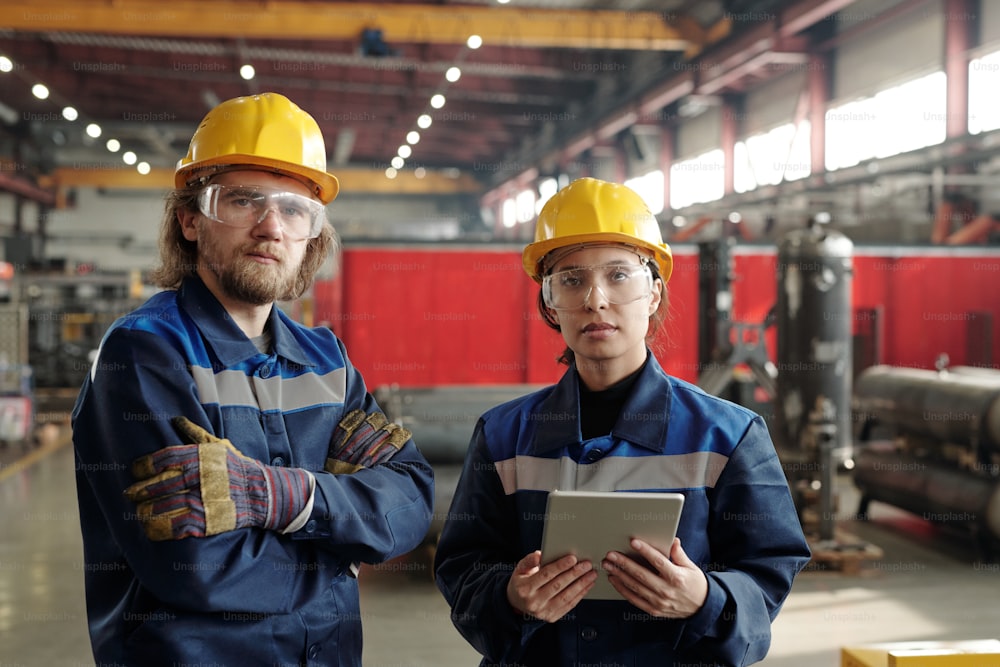 Two young serious engineers in protective workwear looking at you while standing in front of camera, female worker using digital tablet