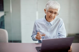 Mature businesswoman using computer while working in the office and drinking a glass of water.