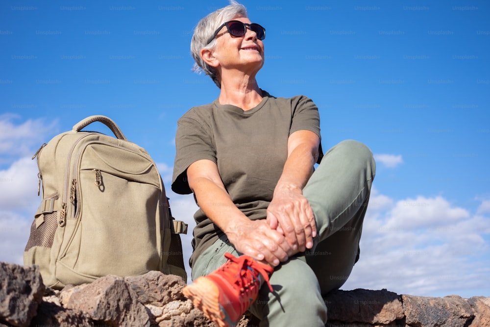 Smiling senior woman in outdoor excursion sitting on a stones wall looking up. Mature active woman sitting close to her backpack enjoying freedom and sunny day