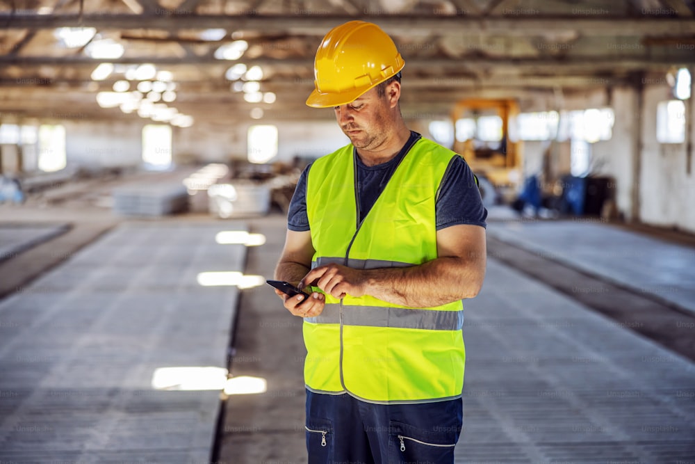 Trabajador de cuello azul trabajador parado en el sitio de construcción y usando un teléfono inteligente para pedir más materiales para la construcción.