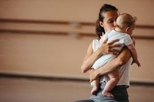 Young loving mother kissing her baby while holding him during exercise class in health club. Copy space.