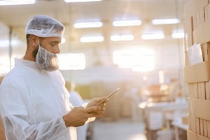 A supervisor with a hairnet and beard net in a sterile white uniform is standing next to a pile of boxes and checking them. He holds the tablet in hand and looking at it.