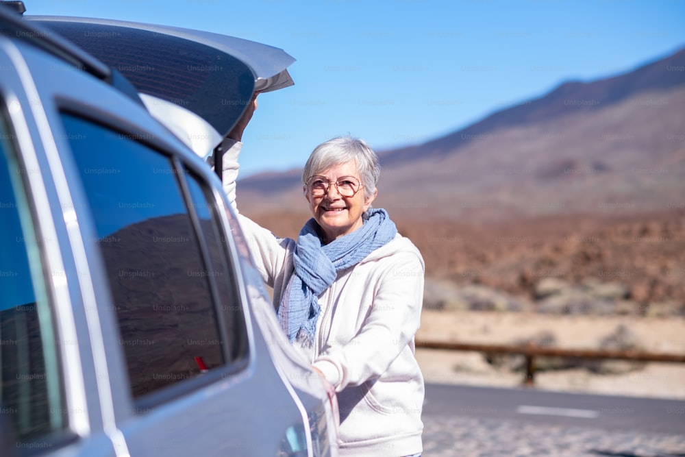 Smiling mature senior woman in outdoors mountain excursion  with automobile opening hood of the car