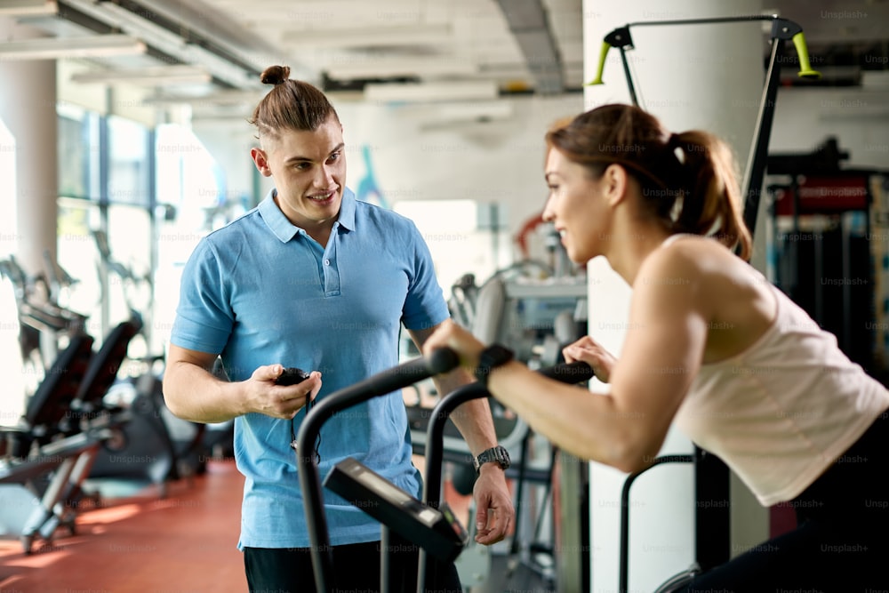 Junger Trainer misst die Zeit auf der Stoppuhr, während eine athletische Frau während des Sporttrainings in einem Fitnessstudio ein Trainingsgerät benutzt.