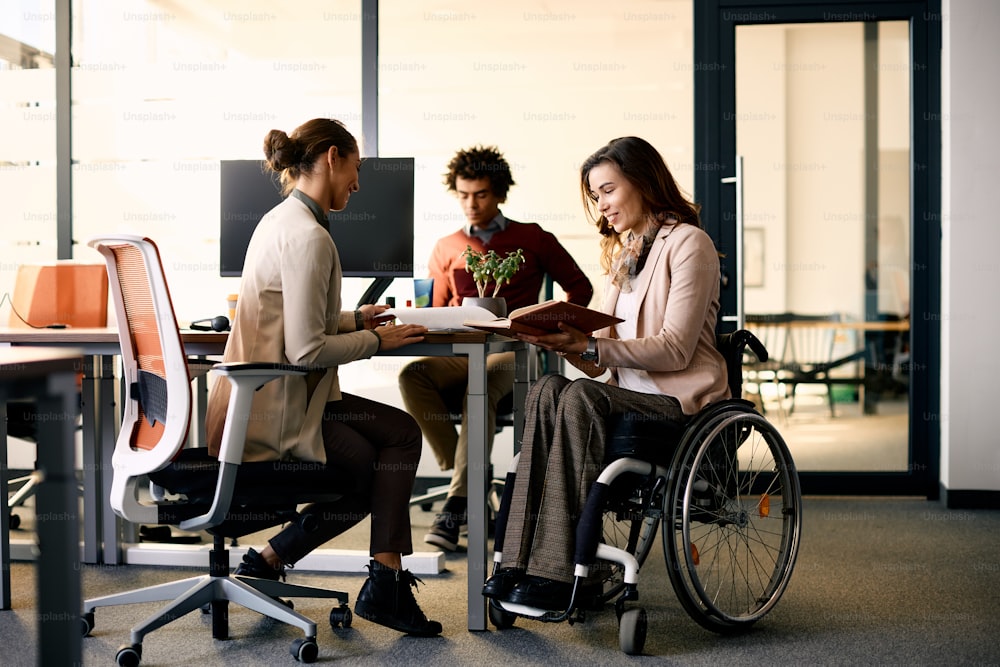 Glückliche Geschäftsfrau mit Behinderung und ihre Kollegin kooperieren bei der Arbeit an Papierkram im Büro.