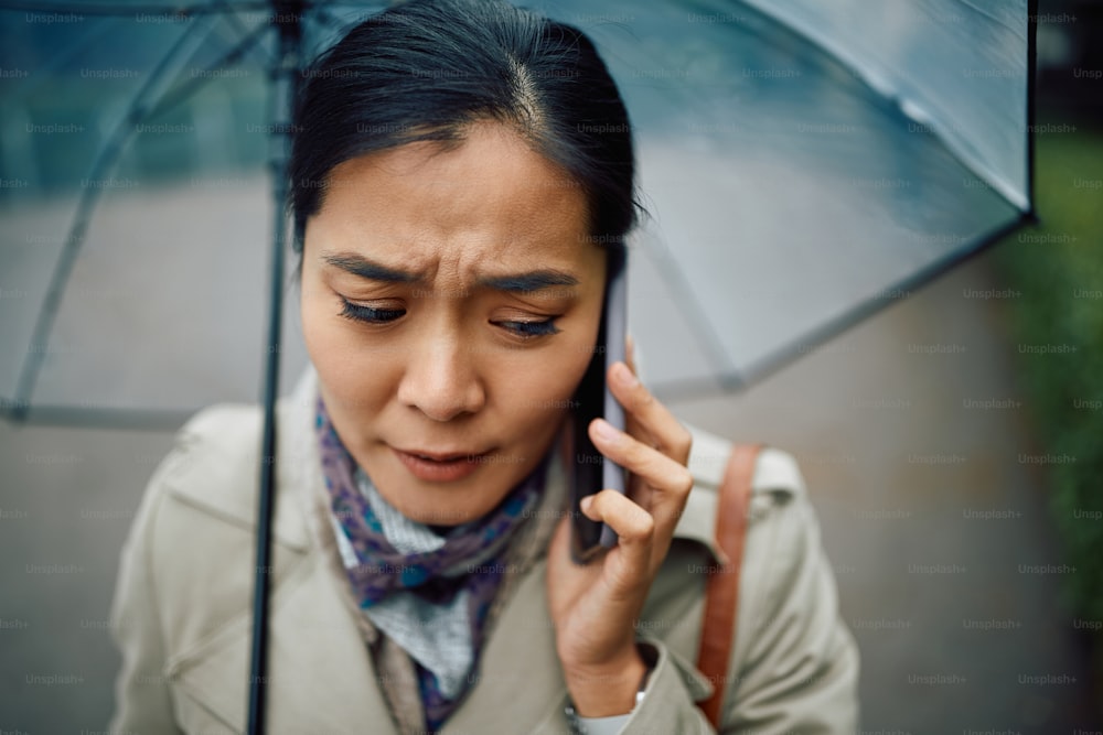 Worried Asian woman receiving bad news during a phone call while walking in the park on rainy day.