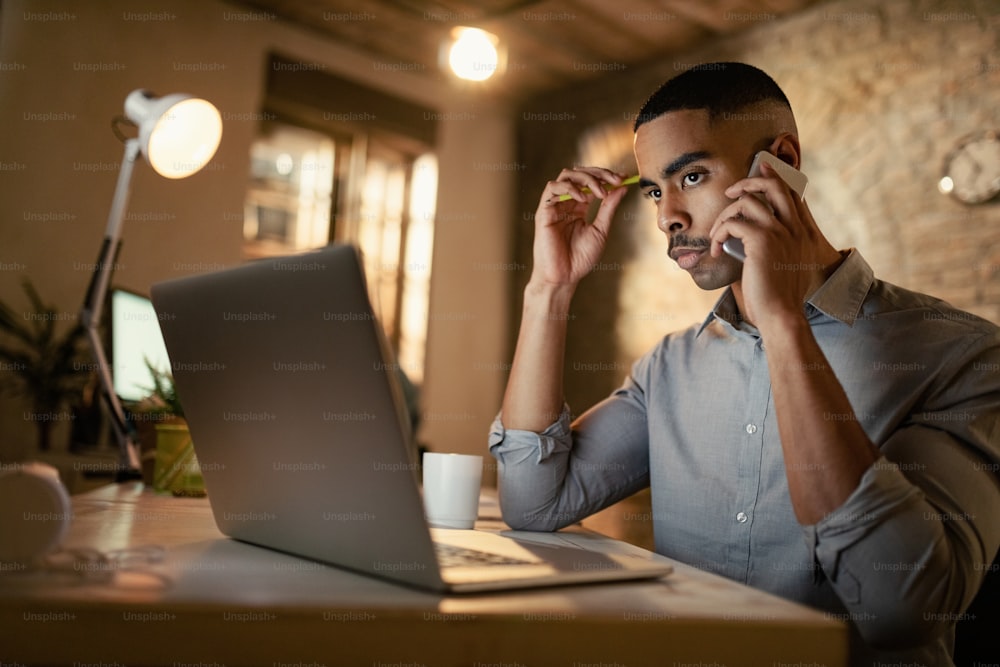 Homme d’affaires afro-américain sérieux faisant un appel téléphonique tout en travaillant sur un ordinateur portable la nuit au bureau.