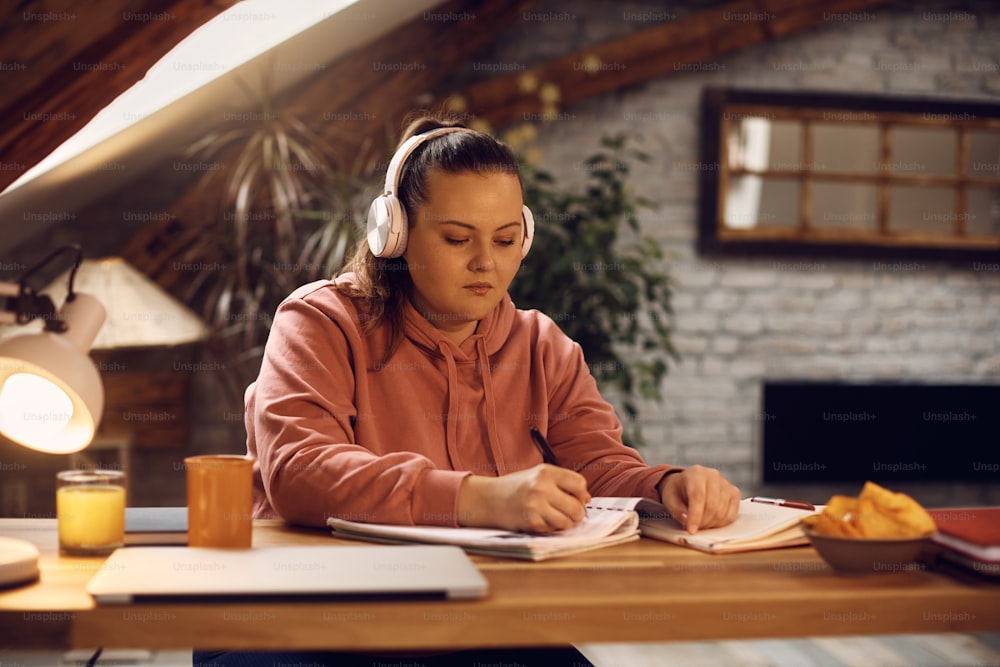 Female college student taking notes and listening music over headphones while studying at home.