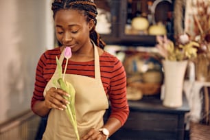 Young African American florist smelling a tulip with eyes closed while working at flower shop.
