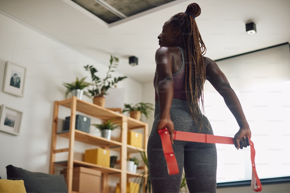Back view of black female athlete using power band while exercising at home.