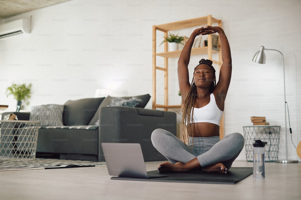 Afroamerikanische Sportlerin, die Laptop benutzt, während sie Yoga praktiziert und sich im Wohnzimmer dehnt.