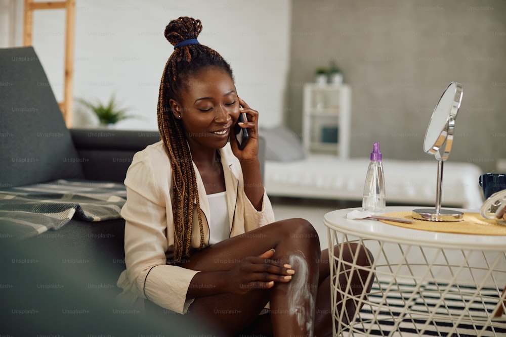 Happy black woman applying body lotion while using mobile phone and talking to someone at home.