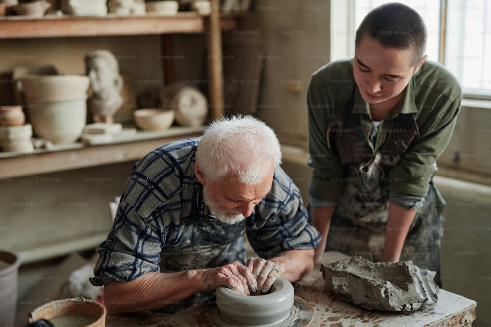 Senior potter sculpting a cup from clay pot with his assistant watching for the process in studio
