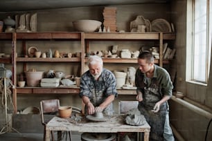 Senior man making earthenware dish on pottery wheel with woman giving him the piece of clay, they working in studio