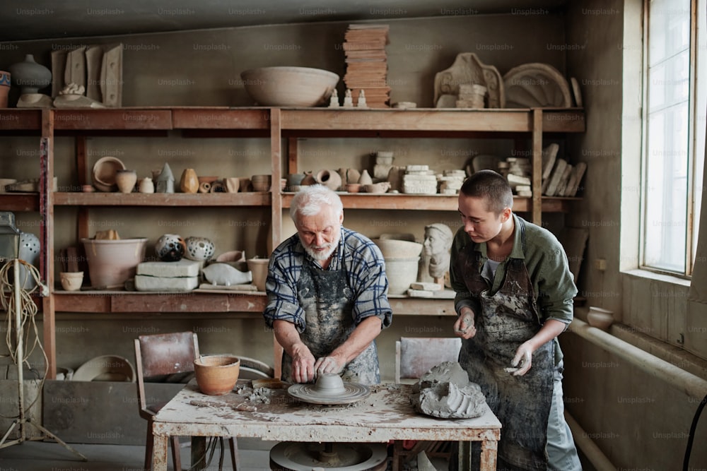 Senior man making earthenware dish on pottery wheel with woman giving him the piece of clay, they working in studio