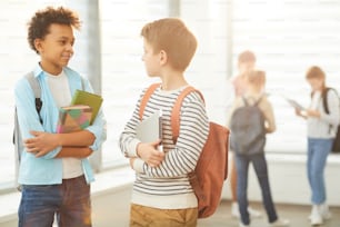 Plano medio horizontal de dos amigos adolescentes de pie en el pasillo de la escuela con mochilas y libros de texto charlando, espacio de copia