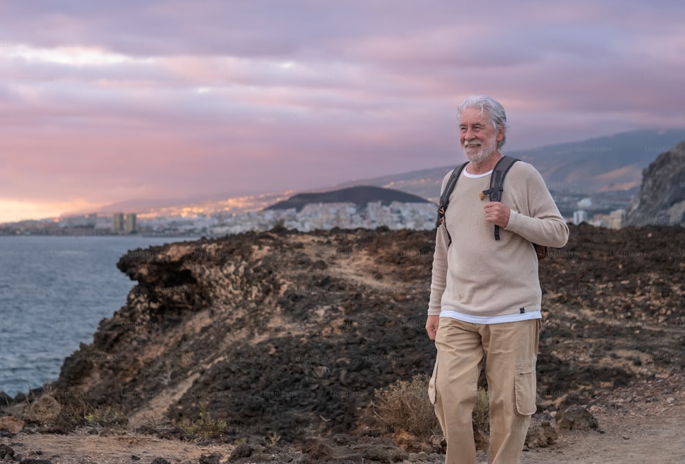 Adult caucasian gray haired senior man walking in countryside close to the sea wearing backpack looking at the horizon enjoying nature. Freedom and vacation concept. Cloudy sky. Copy space