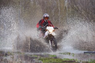 Piloto de motocross compitiendo en madera inundada y salpicando un gran charco a alta velocidad