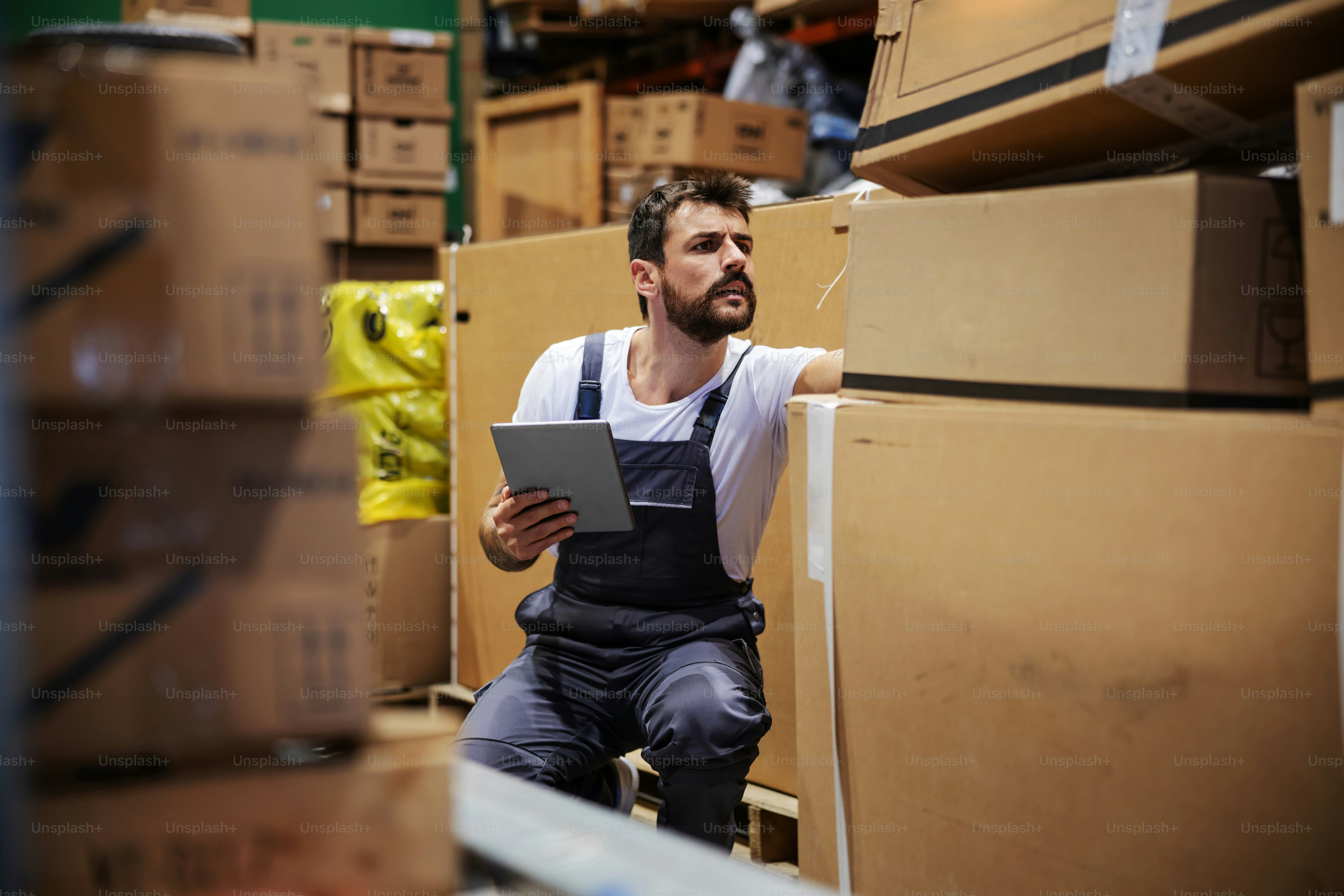 Bearded tattooed hardworking blue collar worker in overalls crouching in storehouse, using tablet and checking on inventory.