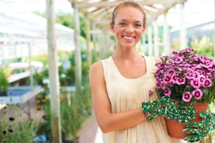 An attractive young woman holding a pot of purple flowers in a garden centre while looking at the camera