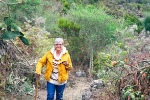 Smiling senior woman outdoor on mountain excursion wearing winter sweater and yellow jacket. Relaxed elderly woman leaning at walking cane enjoying freedom and nature