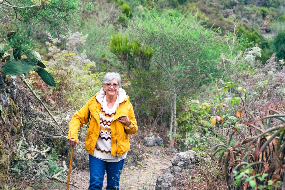 Smiling senior woman outdoor on mountain excursion wearing winter sweater and yellow jacket. Relaxed elderly woman leaning at walking cane enjoying freedom and nature