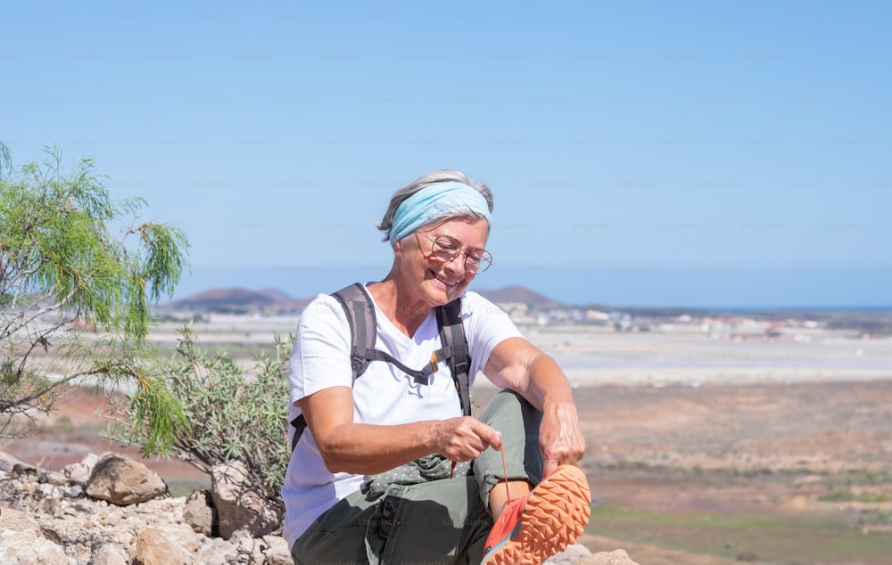 Donna anziana attiva felice con zaino che fa escursioni all'aperto in montagna seduta allacciandosi le scarpe godendo di uno stile di vita sano e di una giornata di sole. Vista panoramica sul mare e sulla montagna sullo sfondo