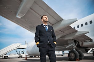 Experienced airplane captain placing hands in his pockets while watching calmly ahead of himself on runway