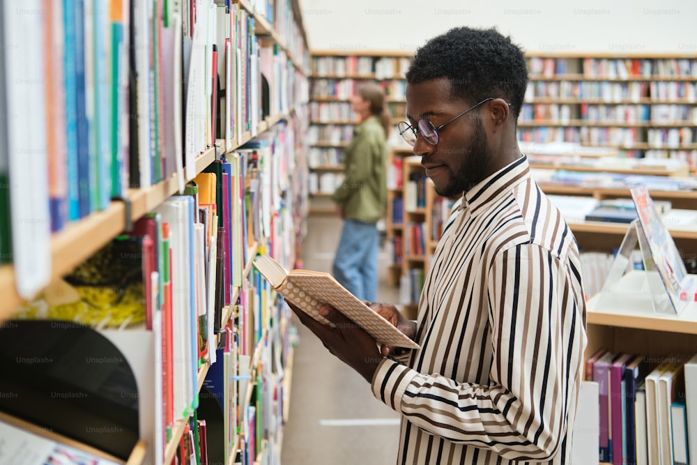 Estudante africano em pé na frente das prateleiras e lendo um livro na biblioteca