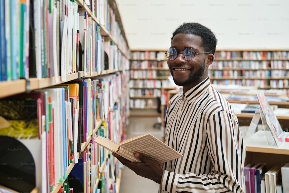 Retrato do jovem africano em óculos sorrindo para a câmera enquanto está em pé na frente da estante th na biblioteca